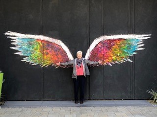 Pugsley posing in front of a wall with colorful wings painted on it. 