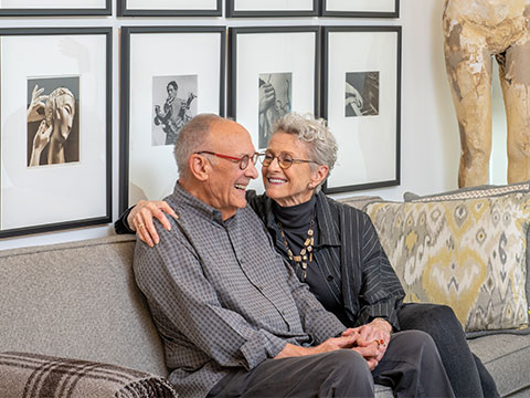 Senior couple holding hands while sitting on their couch inside of their apartment.