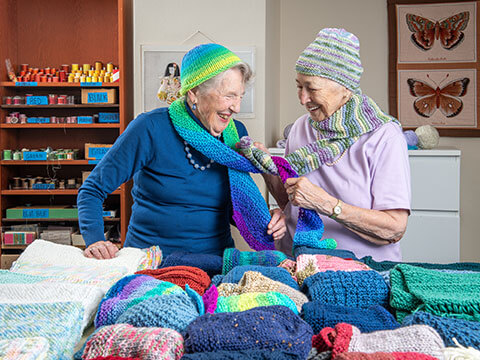 Two senior women laughing while wearing a crocheted hat and scarf.